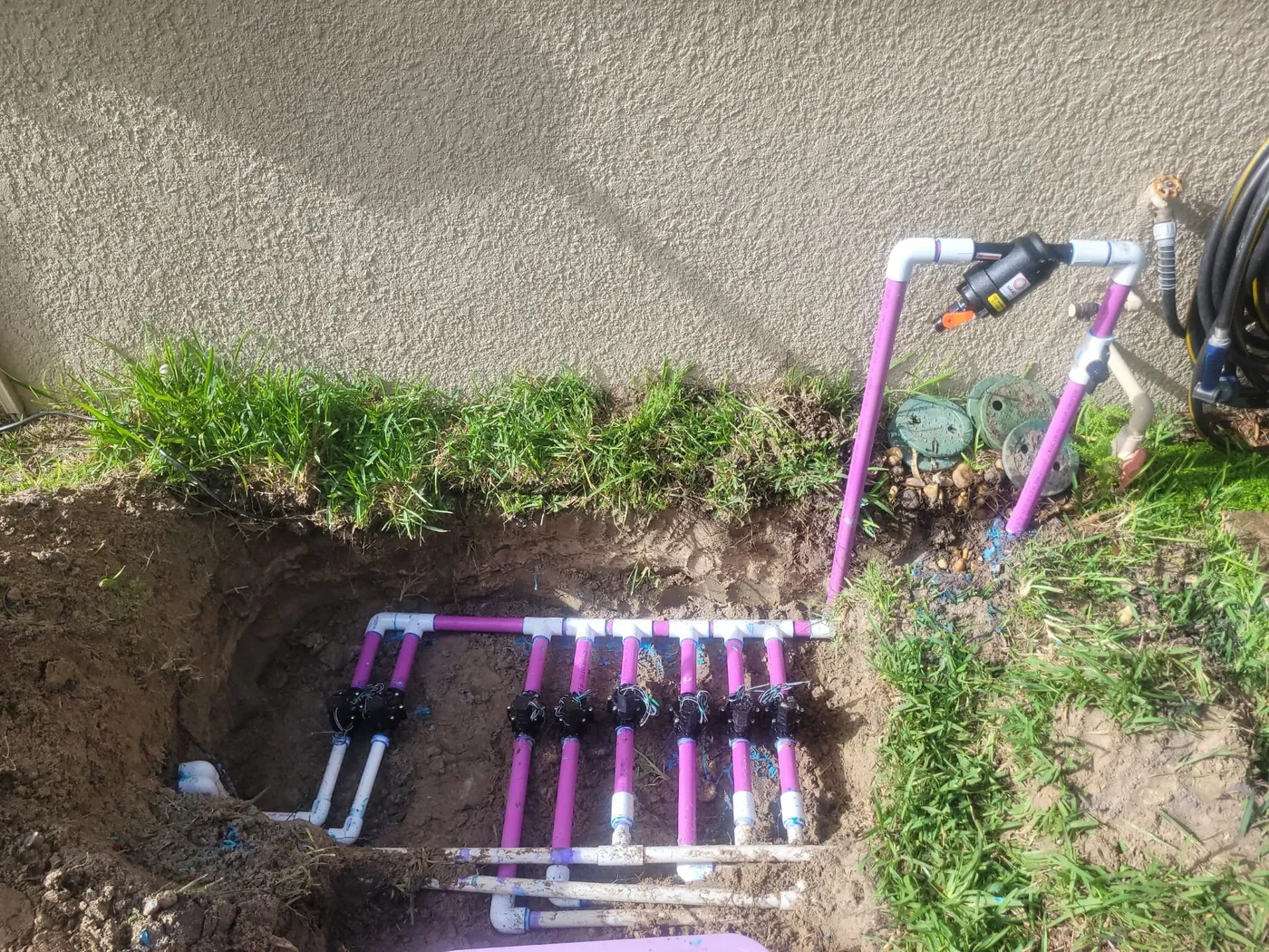 Sprinkler valve manifold with PVC lines laid in an open trench beside a house wall